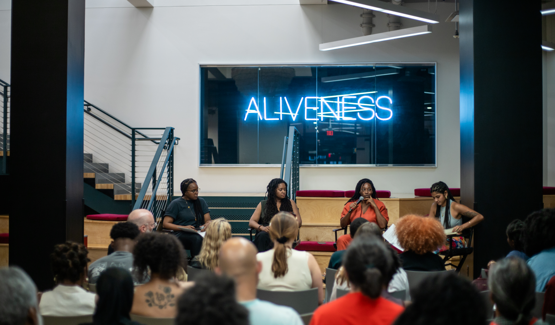 Interior image of Express Newark, a group of panelists are seated behind a blue neon sign that reads "ALIVENESS"